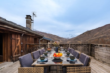 A wooden deck with a dining table set for six, surrounded by wicker chairs, with a mountain backdrop.. Chalet “Le petit Paradis” in the heart of the 3 valleys!