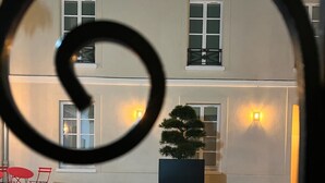 A courtyard with a cobblestone floor, a potted plant, and a building with multiple windows and wall-mounted lights. - Appartement au Coeur de Ville (Sens)
