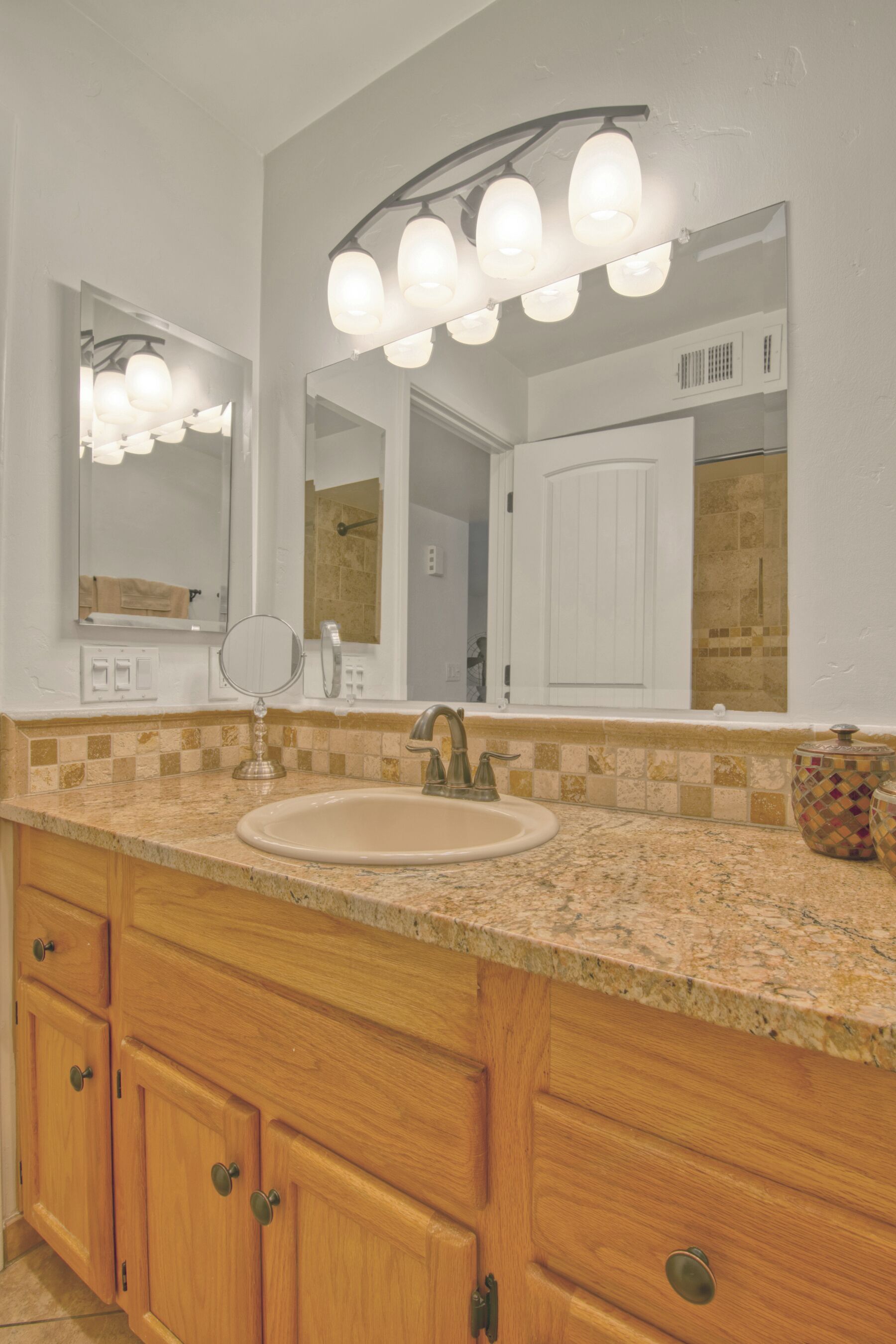 Un baño con encimera de granito, lavamanos, armarios de madera y espejo con luces arriba.