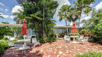 A patio with outdoor furniture and umbrellas, surrounded by lush greenery and palm trees.