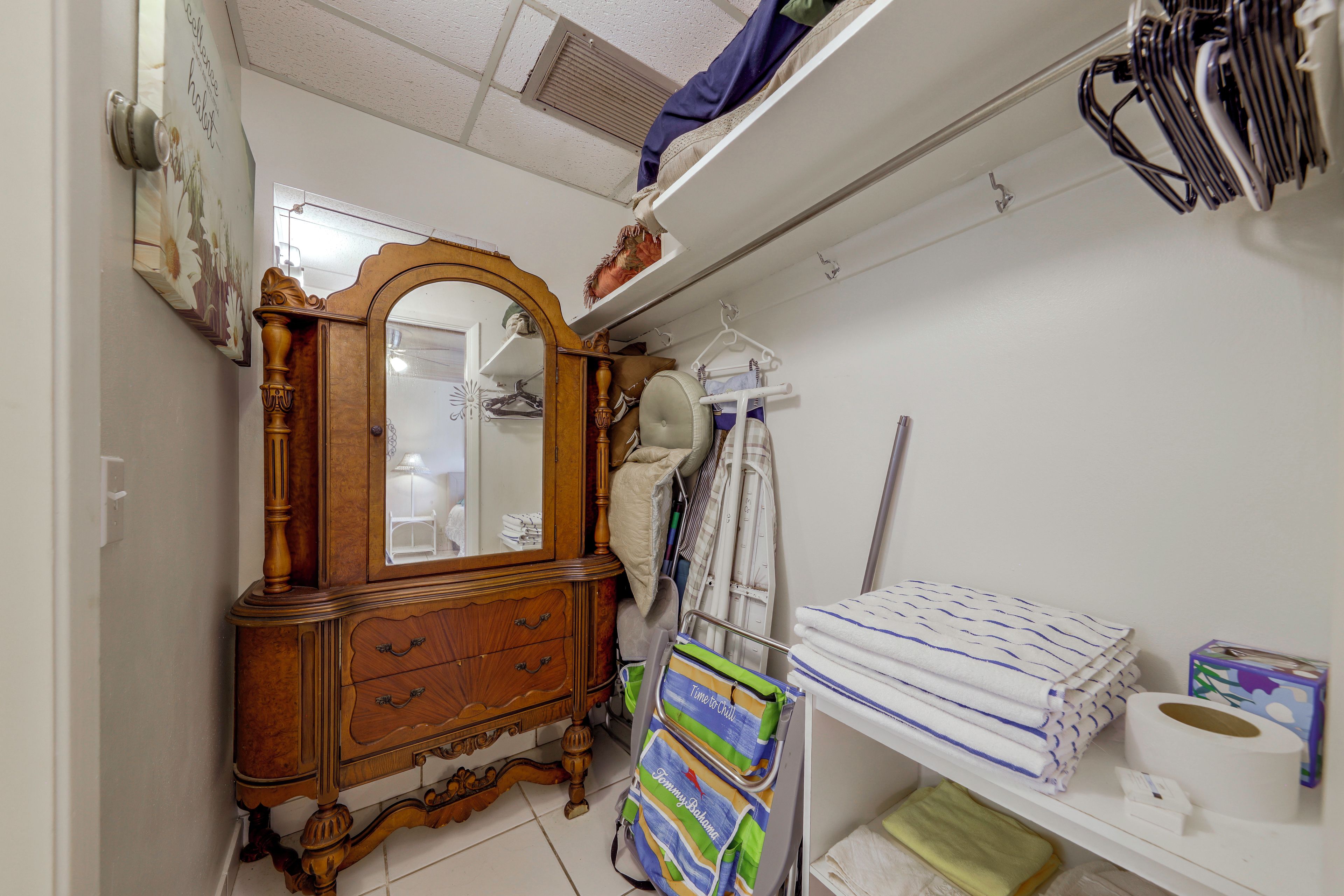 A room with a wooden dresser, a mirror, a hanging rack with clothes, and a shelf with towels.