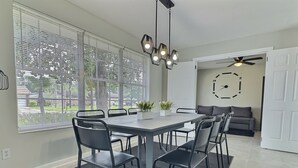Un moderno comedor con una mesa rectangular, sillas negras y un tapete negro. Se ve una ventana con persianas, un ventilador de techo y un reloj en la pared.