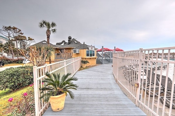 A wooden boardwalk leading to a building with a palm tree and potted plants.
