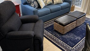 Uma sala de estar com um sofá azul, uma poltrona escura e um banquinho de vime. Há um tapete estampado no chão e um quadro de uma cena de praia na parede.