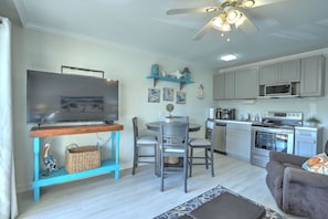 A modern living room with a flat-screen TV, a dining table with chairs, a kitchen with white cabinets and appliances, and a ceiling fan.
