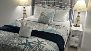 Cama bem arrumada com colcha estampada de estrela-do-mar e corais, duas luminárias de cabeceira e mesa de cabeceira com abajur.