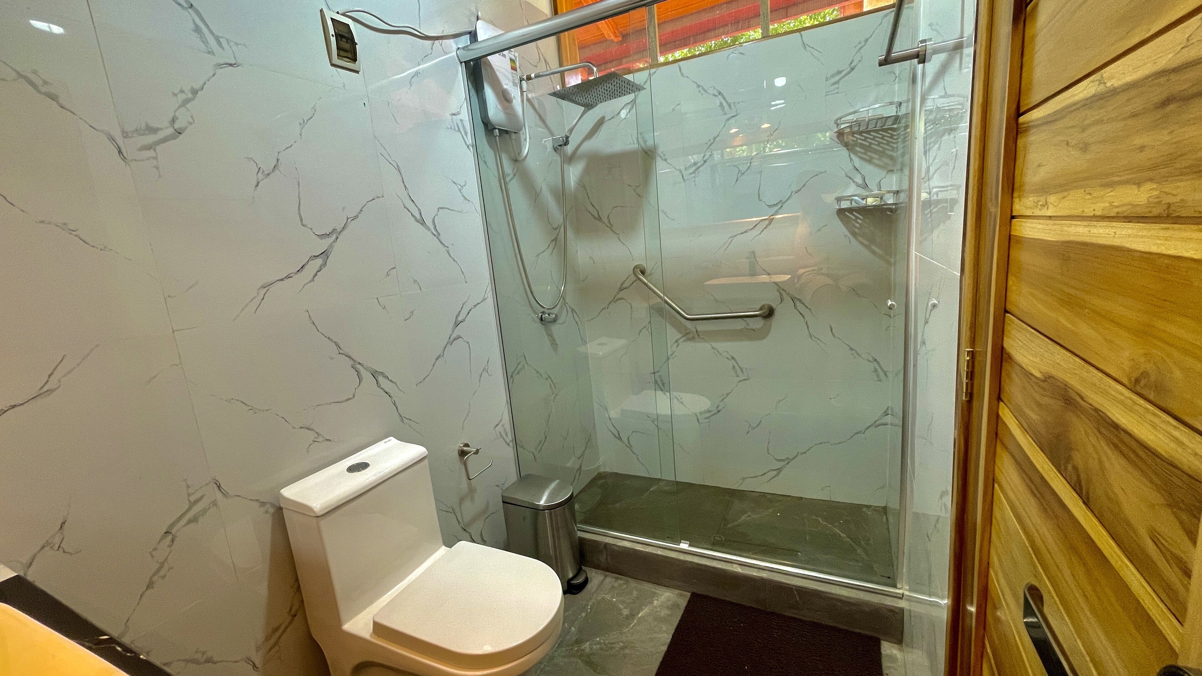 A bathroom with a toilet, a glass-enclosed shower with a handheld showerhead, and a marble-patterned wall.