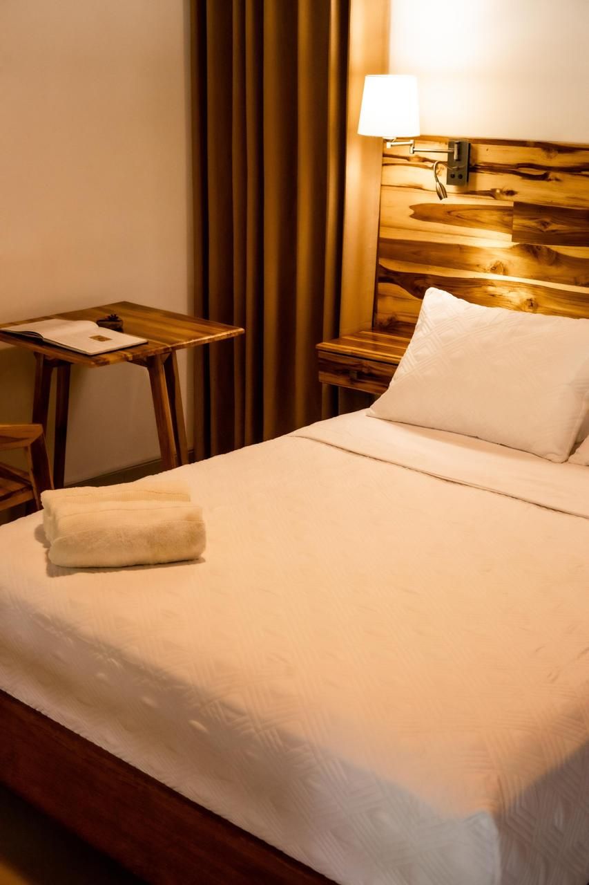 A neatly made bed with a white pillow and a folded towel, a wooden nightstand with a lamp, and a small table with a book and a cup.