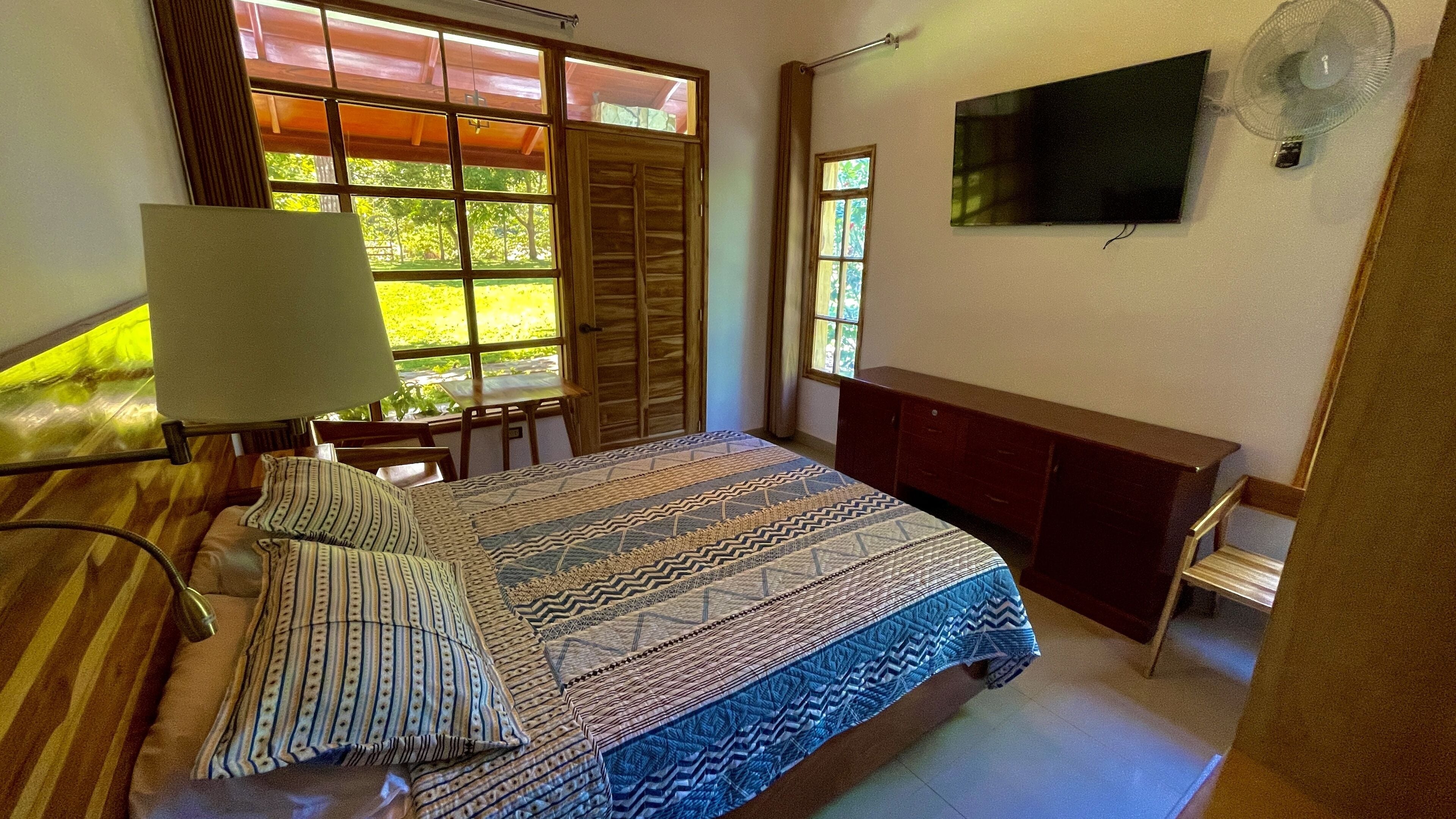 A bedroom with a large bed, a desk, a chair, a TV, and a window with a view of greenery.