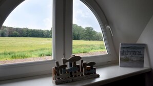 Une pièce dotée d’une grande fenêtre en arc d’où l’on peut admirer un champ verdoyant. Un petit banc en bois est placé sur une petite plateforme devant la fenêtre.