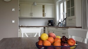 Une cuisine avec des meubles blancs, une table à manger sur laquelle se trouve un saladier rempli de fruits, et une horloge au mur.