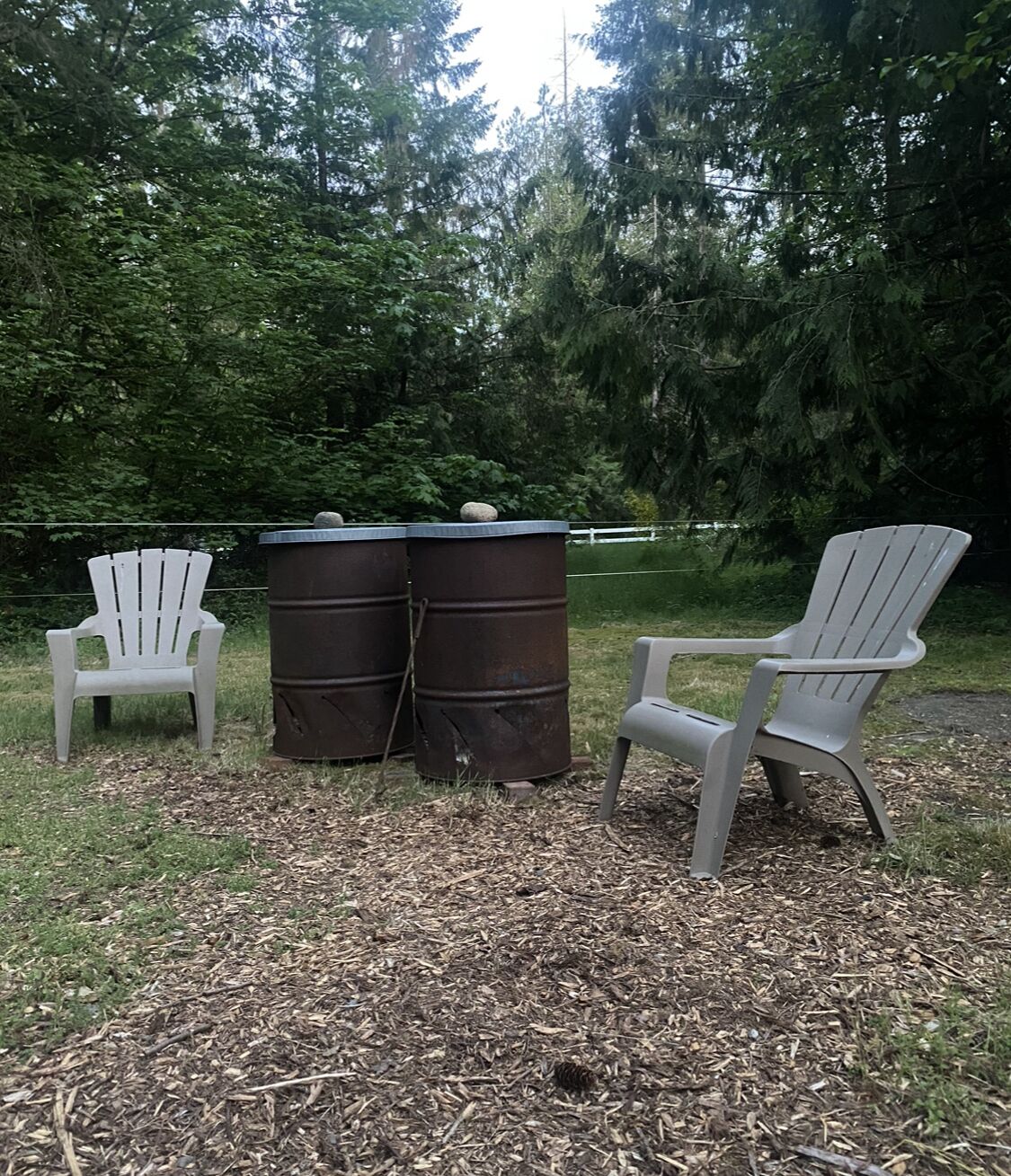 Two Adirondack chairs and two metal barrels in an outdoor setting with a grassy area and trees in the background.