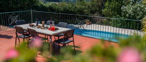A patio with a table set for dining, a swimming pool, and surrounding greenery.