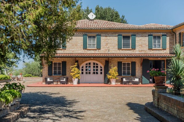 A two-story stone building with a tiled roof, green shutters, and a central entrance with a large arched doorway. There are outdoor seating areas with chairs and tables, and potted plants are placed around the building.