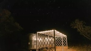 A small, enclosed structure with a lattice roof, surrounded by a dirt ground and sparse vegetation, under a starry night sky.