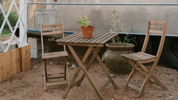 A covered outdoor seating area with a table and two chairs, a potted plant, and a vintage trailer.