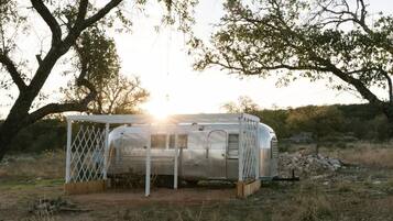 Una caravana antigua con estructura de celosía blanca, ubicada en un entorno natural con árboles y cielo despejado.