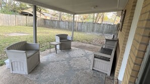 Ein überdachter Patio-Bereich mit Betonboden, einem gemusterten Teppich und zwei weißen Holzstühlen.