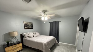 Ein Schlafzimmer mit einem Bett, einem Nachttisch, einer Lampe, einem Deckenventilator, einem Gemälde und einem an der Wand befestigten Fernseher.
