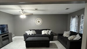 Ein Wohnzimmer mit einem dunkelgrauen Sofa, einem kleinen Hocker, einem Deckenventilator, einer Uhr und einem Fernseher auf einer Kommode.
