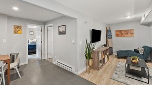 A modern living room with a sofa, a coffee table, and a TV. There is a dining area with a table and chairs. The room has a large window and a door leading to another room. - 8/19 now available Updated 2br Minutes from downtown- backyard & Firepits

 (Colorado Springs)