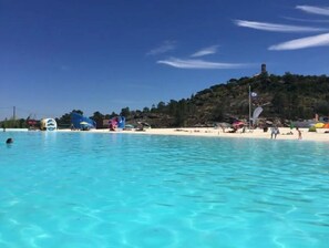 Een strand met helder turquoise water, een zandstrand en een heuvel met een toren in de verte.