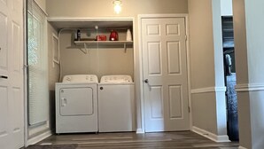 Una sala de lavandería con dos lavadoras blancas, un estante con varios objetos, una puerta y piso de madera.