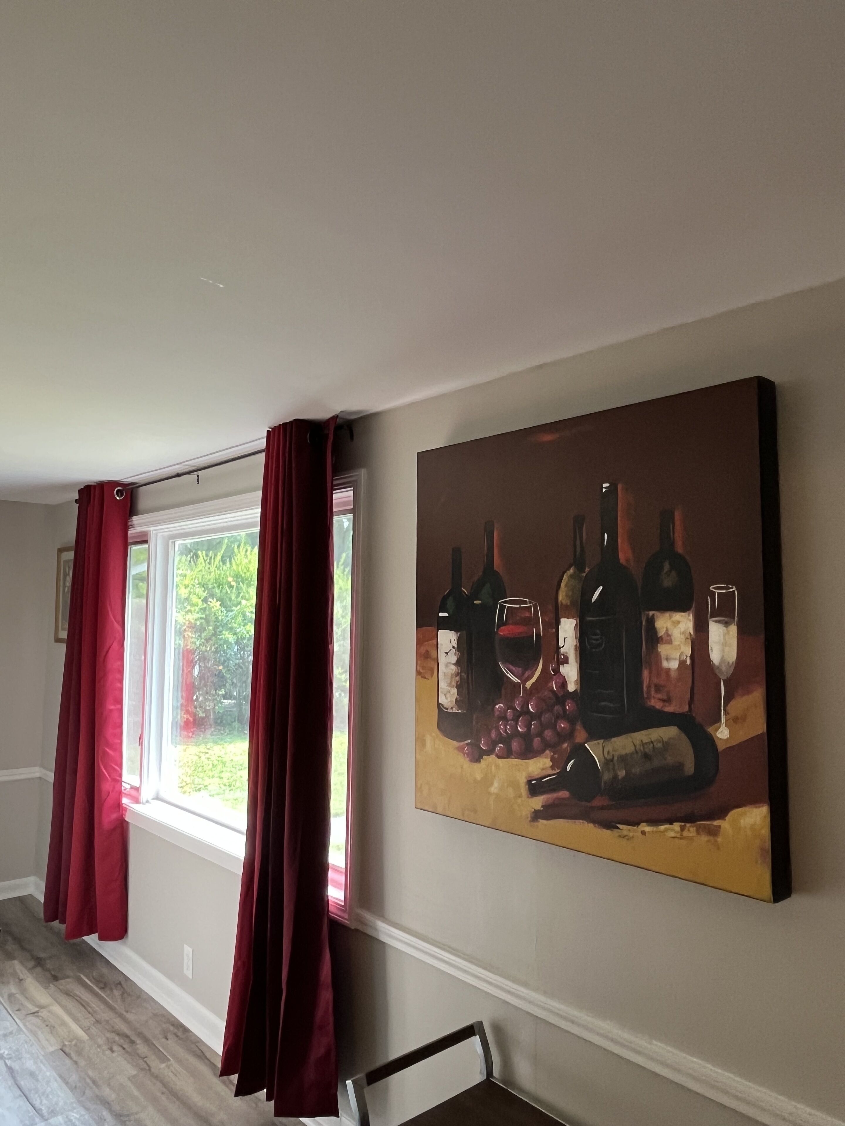 Une pièce avec un grand tableau représentant des bouteilles de vin et des verres sur le mur, une fenêtre avec des rideaux rouges et un plancher en bois.