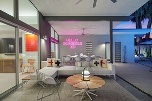 A modern living room with a sofa, chairs, and a coffee table. A neon sign reads "HELLO GORGEOUS XO".