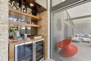 Une cuisine moderne avec un mur en briques, des étagères en bois et une porte en verre donnant sur un coin repas où se trouve une chaise rouge.