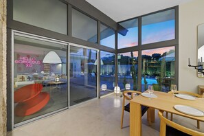 Un espace repas moderne doté d’une grande paroi vitrée, offrant une vue sur la piscine, et agrémenté d’un panneau lumineux au néon.