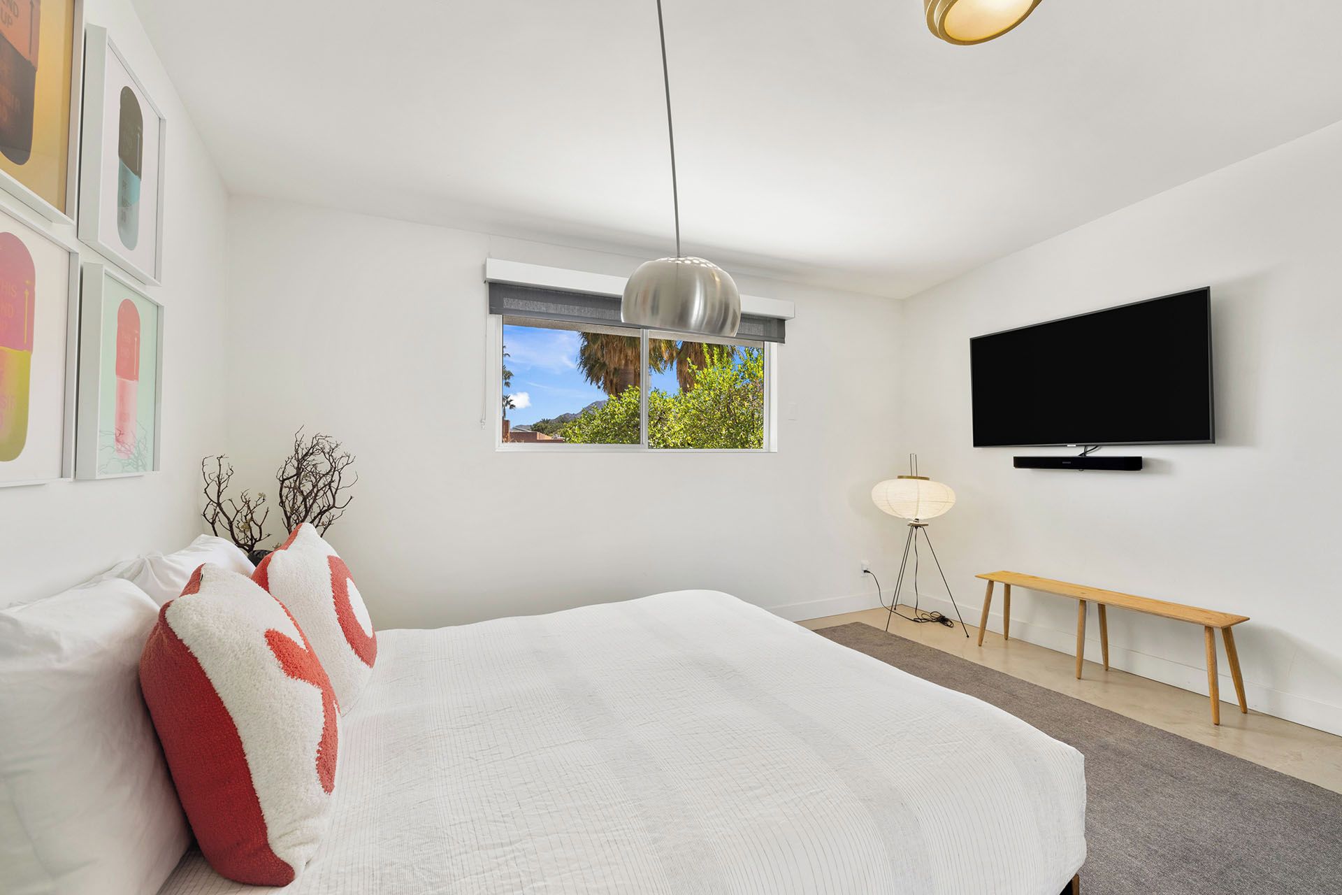 A modern bedroom with a large bed, a TV mounted on the wall, a window with a view of trees, and a bedside lamp.