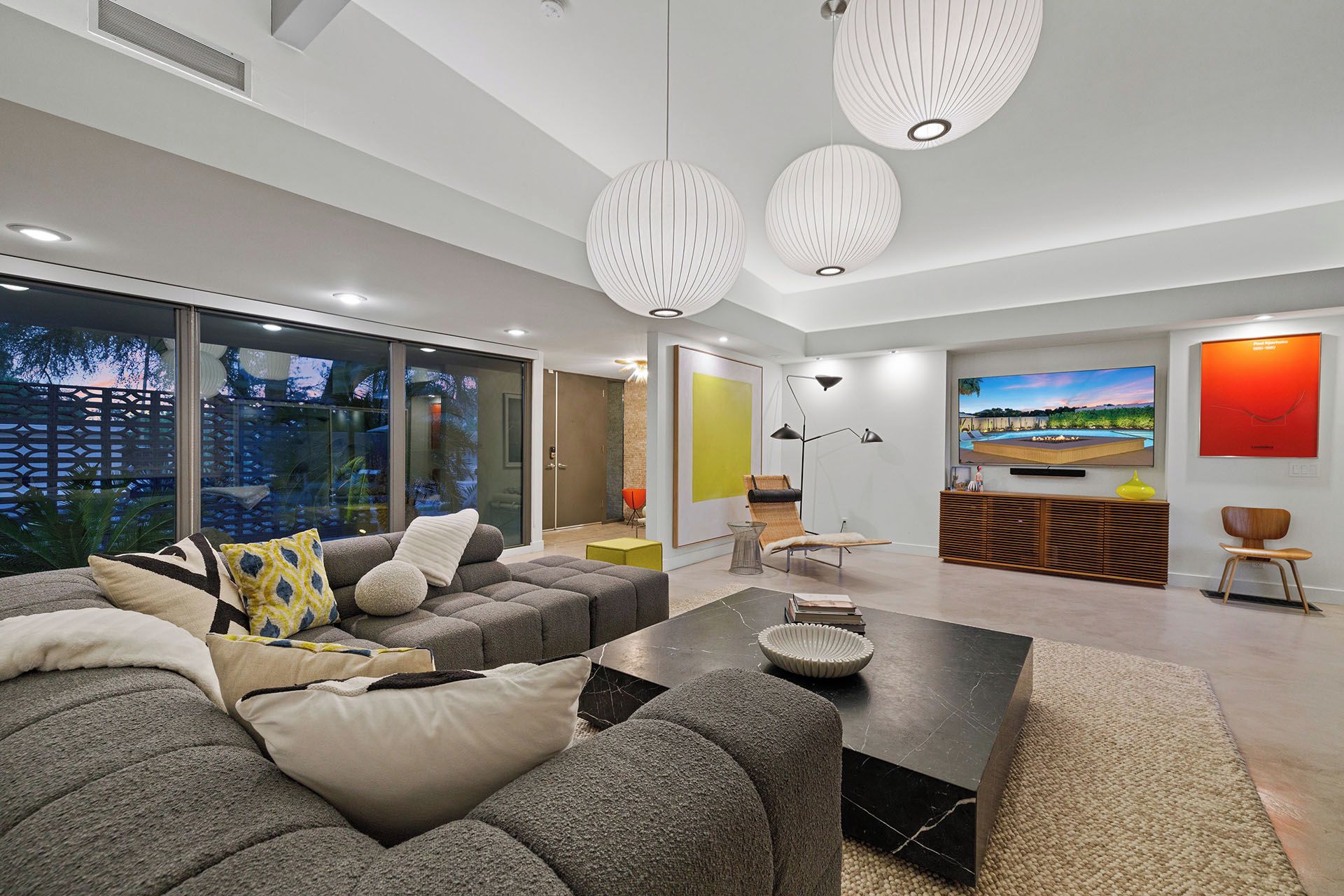 A modern living room with a grey sectional sofa, a coffee table, and a TV mounted on the wall. There are pendant lights, a large window, and a view of the outdoors.