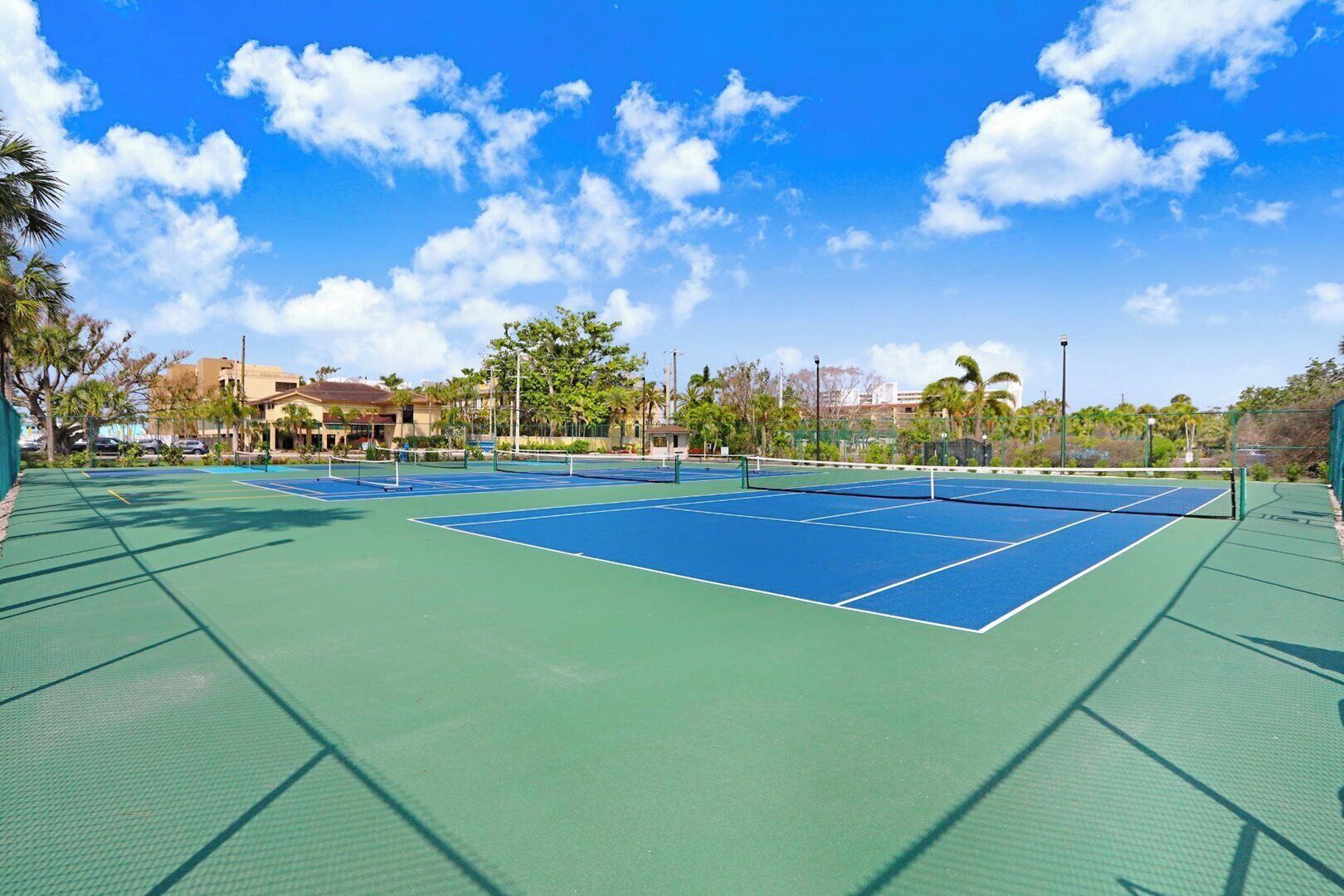 Un campo da tennis all'aperto con superfici verde e blu, circondato da palme e edifici.