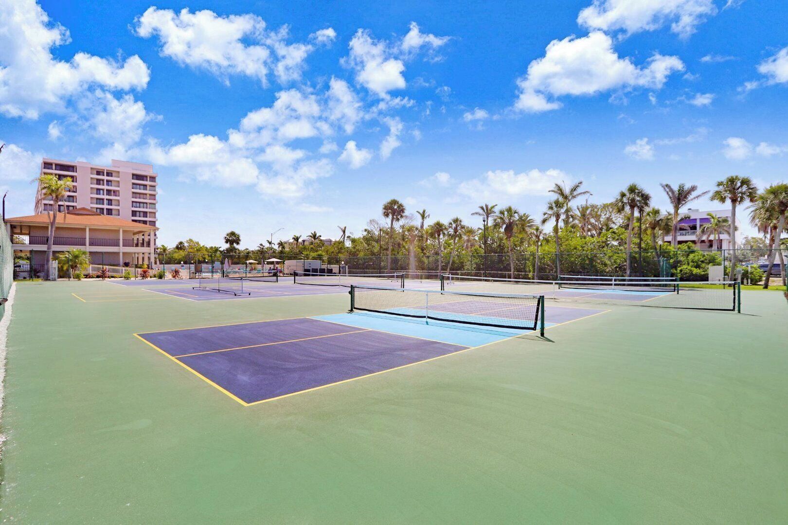 An outdoor tennis court with a net, surrounded by palm trees and a building in the background.