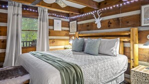 Une chambre dans un chalet en bois, avec un lit, des rideaux, un ventilateur de plafond, des guirlandes lumineuses et une décoration représentant une tête de cerf.