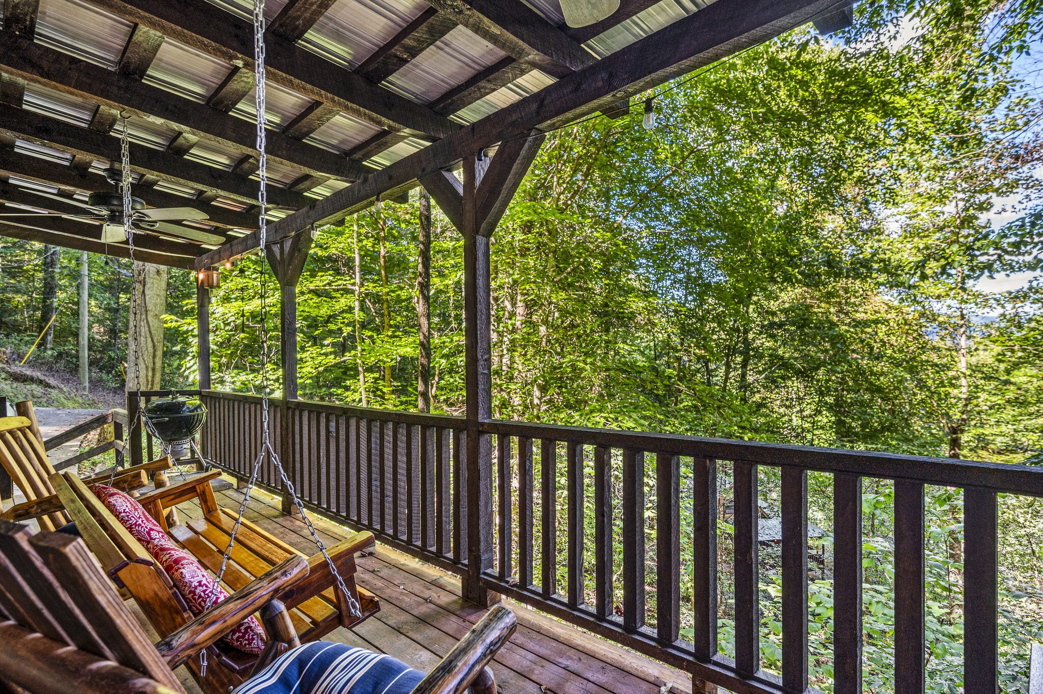 Eine Holzterrasse mit Schaukelstuhl und Grill, mit Blick auf ein bewaldetes Gebiet.