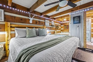 Une chambre confortable dans un chalet en bois, avec un grand lit, des poutres apparentes, des guirlandes lumineuses et un ventilateur de plafond.
