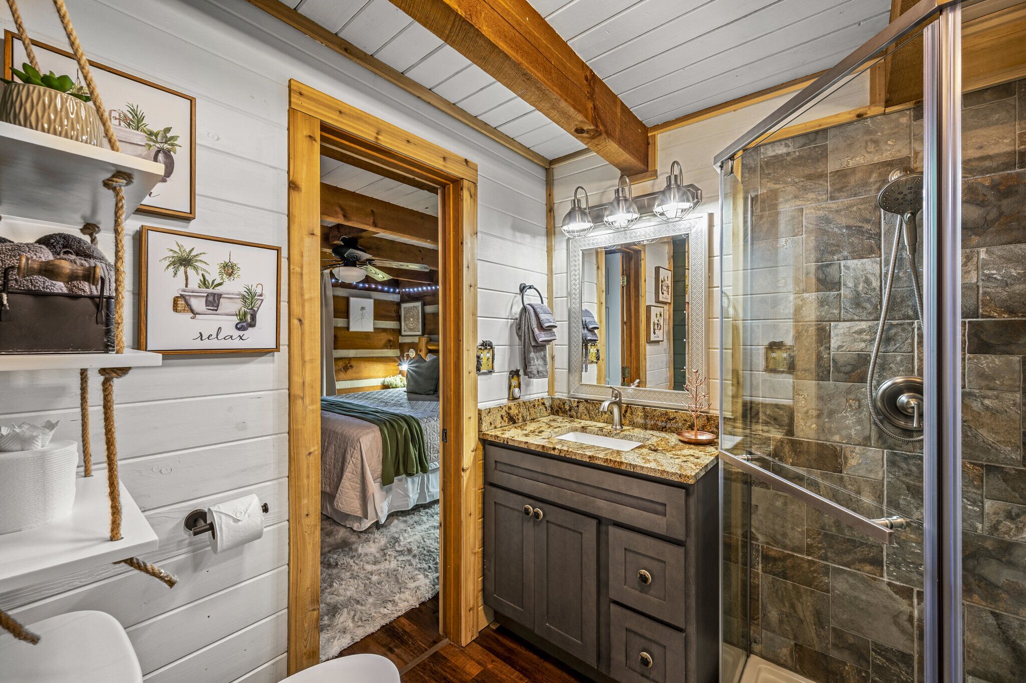 A bathroom with a stone shower, wooden beams, a sink with a granite countertop, and a bed with a green blanket.