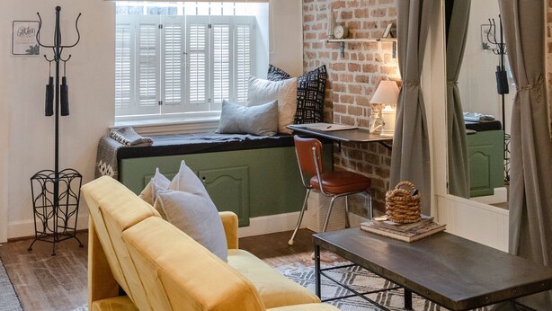 A cozy living room with a yellow sofa, a green armchair, a small table, and a brick wall. There is a window with white shutters and a hanging light fixture. - Historic Garden Studio, Steps from Pulaski Square⎮On-Site Parking (Savannah)