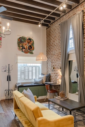 A cozy living room with a yellow sofa, a green armchair, a small table, and a brick wall. There is a window with white shutters and a hanging light fixture. - Historic Garden Studio, Steps from Pulaski Square⎮On-Site Parking (Savannah)