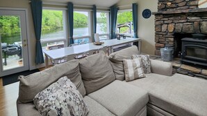 A modern living room with a sectional sofa, a stone fireplace, and a view of a lake through large windows. - SECLUDED WATERFRONT HOME WITH ROOM FOR ALL (Hopkinton)