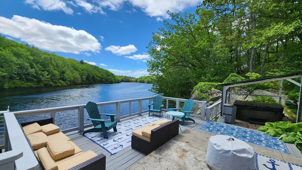 A deck with outdoor seating, a view of a river, and a forested area. - SECLUDED WATERFRONT HOME WITH ROOM FOR ALL (Hopkinton)