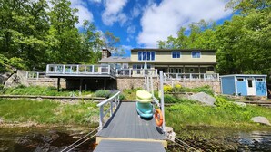 A dock leading to a house with a stone foundation, a covered area with seating, and a small blue shed. - SECLUDED WATERFRONT HOME WITH ROOM FOR ALL (Hopkinton)