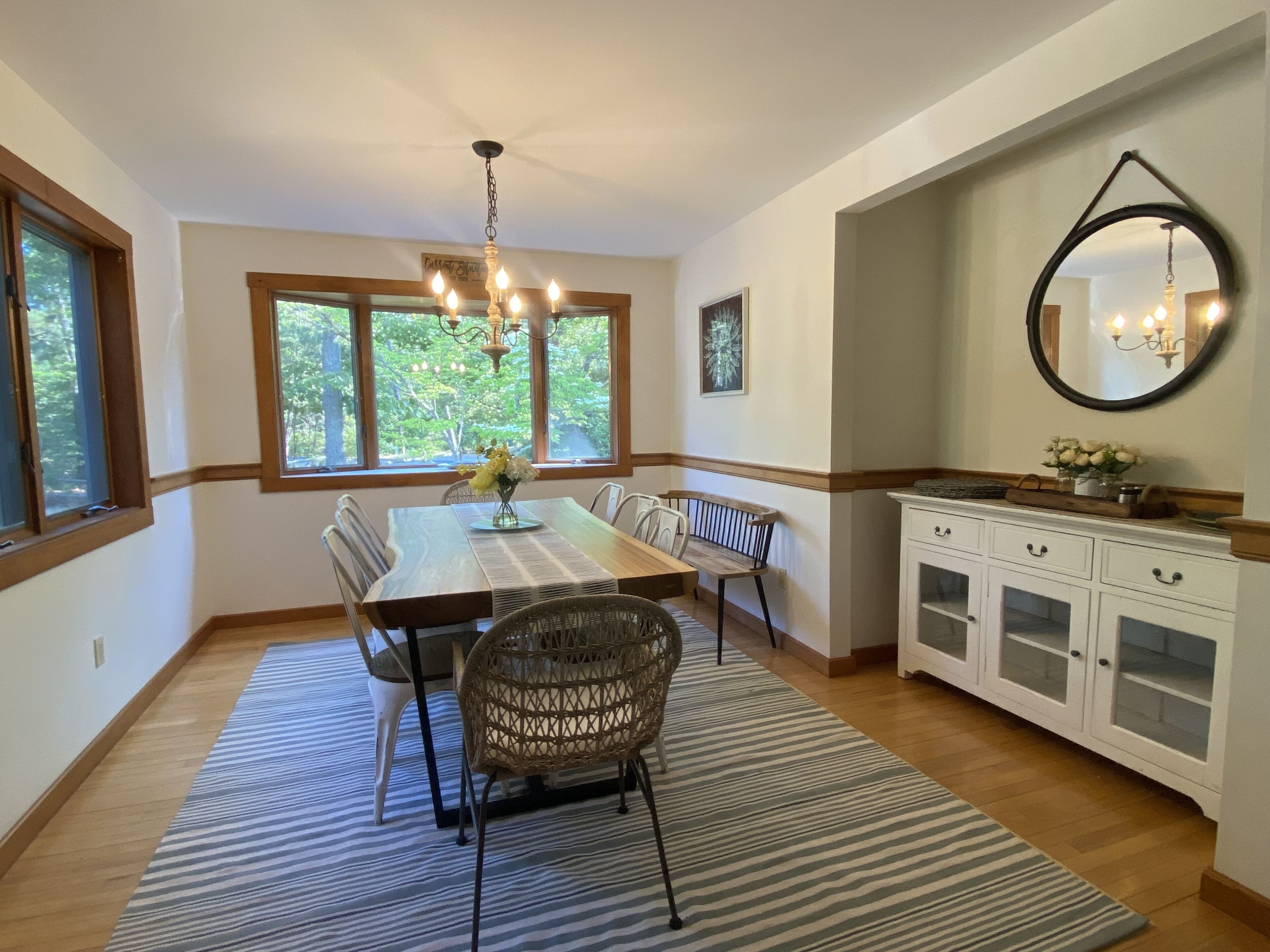 Une salle à manger avec une table en bois, des chaises, un lustre, une fenêtre donnant sur des arbres et un buffet blanc aux portes vitrées.