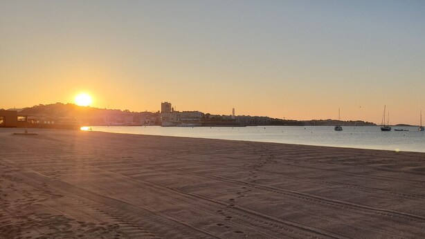 Une plage avec un coucher de soleil, des bateaux et des bâtiments en arrière-plan.