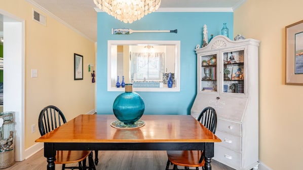 Une salle à manger avec une table en bois, des chaises, un grand vase, un meuble contenant des objets décoratifs et une fenêtre avec des stores.