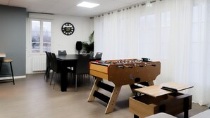 Um quarto com mesa de pebolim, uma mesa de jantar com cadeiras, um relógio na parede e uma janela com cortinas.