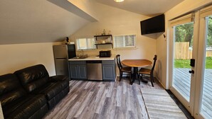 Cocina compacta con refrigerador, microondas y pileta. Hay una mesa de comedor de madera con sillas, un sofá de cuero negro y una puerta corrediza de vidrio hacia el exterior.