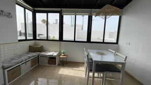 A room with a built-in bench, a dining table with chairs, a small side table, and large windows with a view of the cityscape. - Ocean Tabaiba • Pool • 30M to the Beach • LVH (Puerto del Carmen)